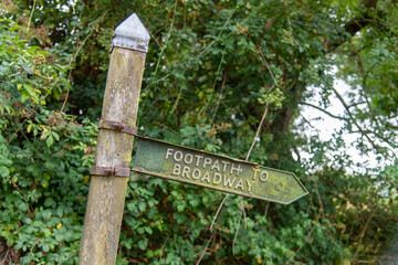 Close up view of a crooked standing wooden pole with sign indicating the footpath to Broadway, Cotswolds, UK in the middle of green trees and bushes and sign cover with green algae
