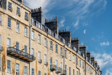 Low angle view of a long bending row of Georgian townhouses with characteristic roofs and chimneys in Bath, Sommerset, UK against a blue sky with feather clouds and half moon