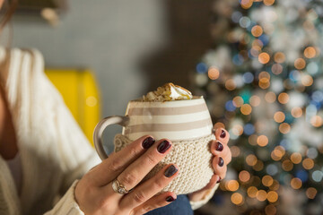 Morning coffee with whipped cream. winter composition. Hand with a cup of Cappuccino or latte coffee on christmas lights background.mug of hot cocoa in the hands of a girl with a dark red manicure