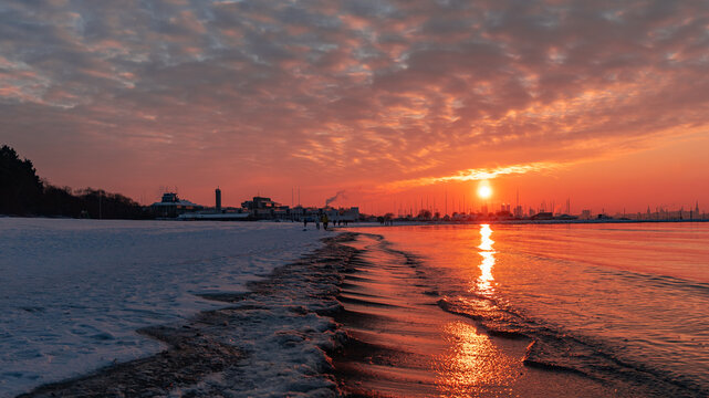 Beautiful sunset on the coast in winter. White snow lies on the embankment. Winter city beach. Amazing clouds sky horizon. Baltic Sea. Tallinn silhouette. Bright golden orange pink purple tones. 