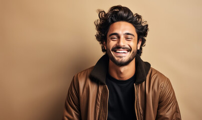 Joyful young man in his 20s with a charming smile wearing a brown leather jacket over a black sweater, studio portrait with a warm beige background