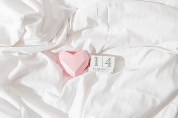 Valentine's Day concept, pink paper valentine heart and 14 february holiday date on wooden calendar in bed on on  white crumpled sheets. Minimal style flat lay photo, top view, copyspace, pastel