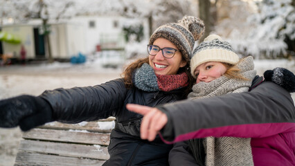 Joyful Winter Moments: Two Young Women in Wool Hats and Scarfs Pointing and Laughing on Snowy Bench