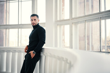 Handsome young man in black turtleneck leans on balcony railings, with large windows in the background - 687952604