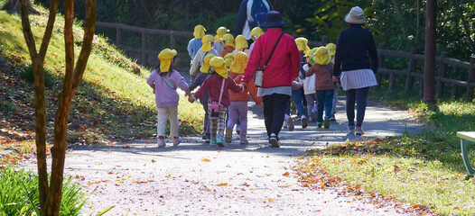 秋の公園で散歩している保育園の園児と先生の姿 - obrazy, fototapety, plakaty