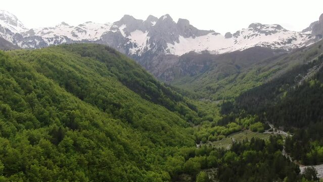 Drone view in Albania flying in the alps showing green forest on a valley surrounded by mountain with snowy peaks in Valbon&euml;