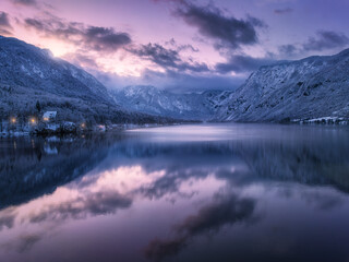Aerial view of lake, snowy alpine mountains, pine trees in snow, reflection in water, purple sky...