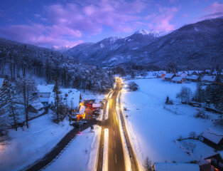 Aerial view of road, snowy mountains, street lights, forest in snow, purple sky at night in winter....