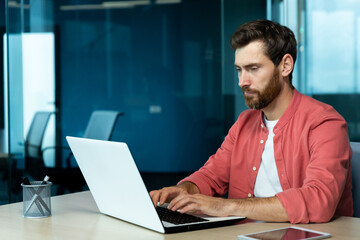 Serious concentrated thinking businessman working with laptop inside office at workplace, man typing on keyboard, preparing online presentation and financial accounting report.