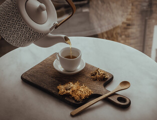 Pouring Chinese chrysanthemum flower tea from Ceramic teapot into a white teacup on wooden cutting...