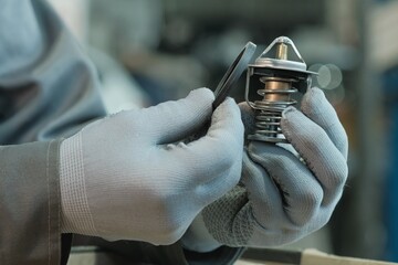Car maintenance at a car service center. An auto mechanic holds a thermostat. The specialist replaced and installed a new seal on the thermostat. - obrazy, fototapety, plakaty