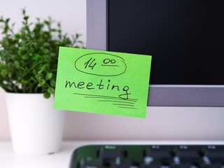 A green paper notes with the reminder 14-00 Meeting on it sticked on to a monitor at an office workplace.