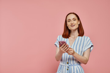 cheerful stylish woman in striped dress holding smartphone and looking at camera on pink backdrop
