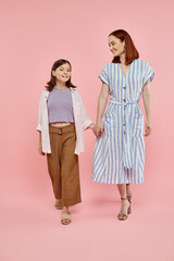 full length of cheerful and stylish woman with preteen daughter holding hands and walking on pink
