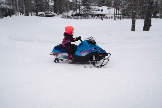 A child in a pink helmet and dark winter clothing rides a blue snowmobile on a snowy trail, with a backdrop of pine trees and overcast skies.