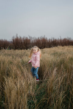 A female child in a pink puffer jacket enjoys a carefree walk through a vast field of tall dry grass, signaling the late autumn vibe.