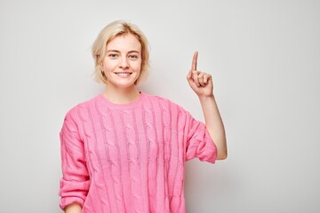 Young blond woman in pink sweater pointing up finger, demonstrating empty space for product or text...