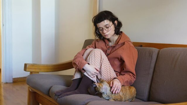 Lonely depressed teenager sitting on sofa, stroking beloved adorable breed cat Devon Rex. Teen girl in need of comfort, emotional support, empathy, contact. Fluffy pet performing antistress function