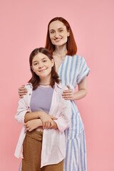 cheerful and stylish woman hugging shoulders of teenage daughter and looking at camera on pink