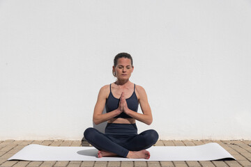 Woman sitting by a white wall on a yoga mat, meditating in the morning sun.