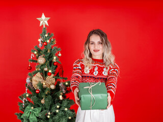 Giving gift box, portrait of smiling beautiful blonde caucasian woman giving gift box. Standing near decorated Christmas tree, wear cozy sweater hold present with ribbon bow. Red background.