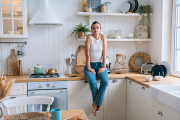 Unhappy brunette girl in casual clothes sitting at kitchen feels loneliness.Disappointed housewife bored at home after divorce. Domestic leisure, sad people. - obrazy, fototapety, plakaty
