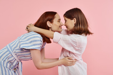 side view of cheerful mother and preteen daughter posing face to face on pink, happy family