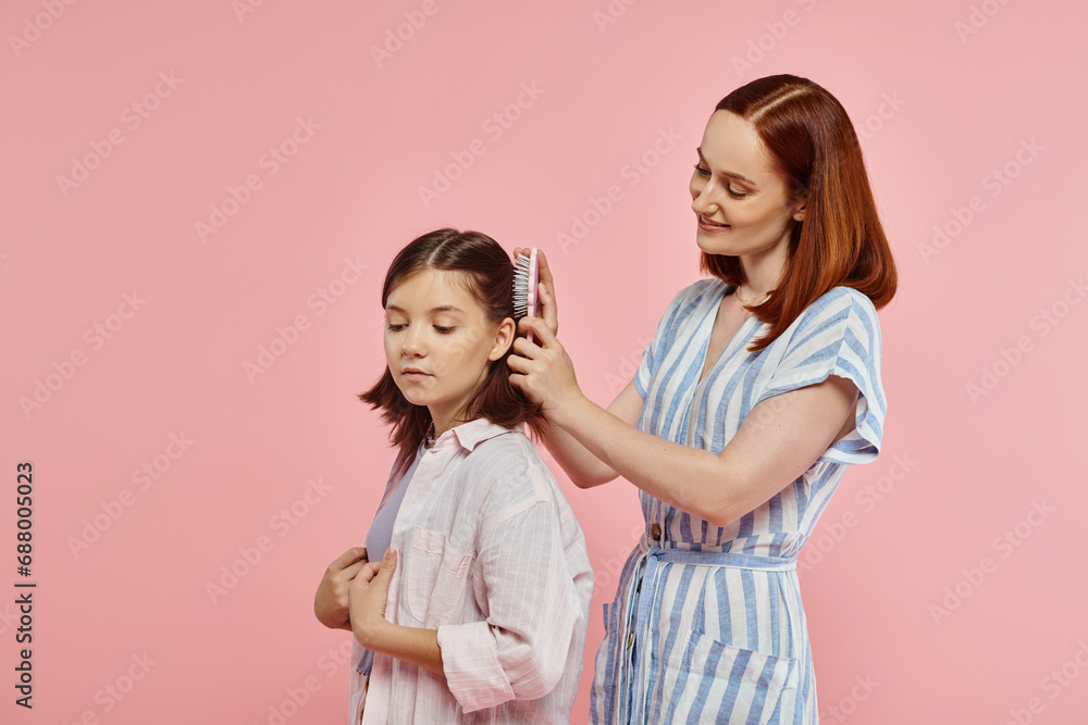 Wall mural smiling mother brushing hair of happy teenage daughter on pink backdrop in studio, care and love