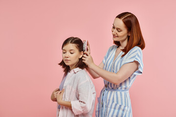 smiling mother brushing hair of happy teenage daughter on pink backdrop in studio, care and love