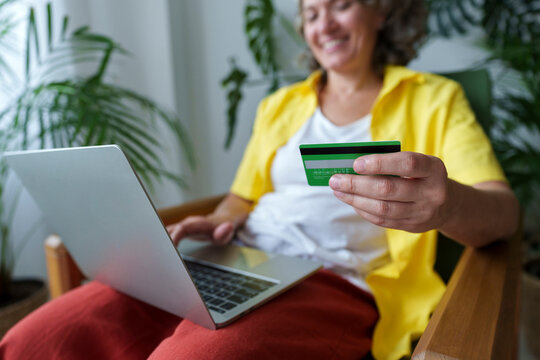 Mature Woman Holding Credit Card Sitting On Armchair Using Laptop At Home