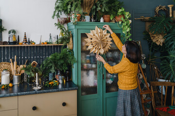 Senior woman hanging paper snowflake on showcase in kitchen at home