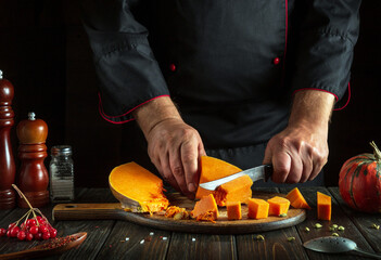 The chef cuts a pumpkin with a knife on a wooden board. Fresh raw diced pumpkin on the kitchen table. Preparing ingredients for a seasonal autumn dish in a restaurant