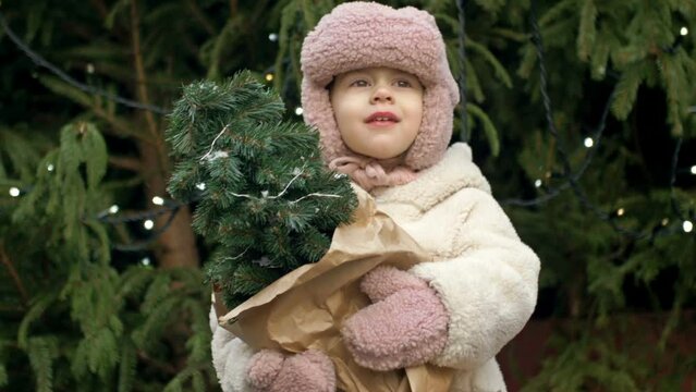 A 2-3 year old girl at the Christmas market holding a small Christmas tree. A child catches the falling snowflakes with her tongue.