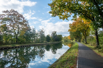 Fototapeta na wymiar This tranquil image captures the essence of an autumn day beside a calm canal. A paved pathway guides the viewer's eye through a scene framed by trees with leaves in shades of green and gold. The