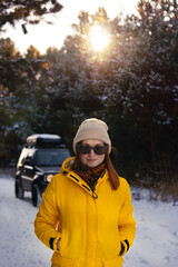 Fototapeta na wymiar Close-up portrait of a young woman in a yellow warm jacket is in a good mood, having fun at sunset on a winter day. Sunglasses. Off-road vehicle and coniferous forest in the background. Vertical photo