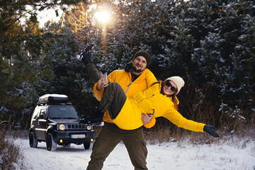 Young couple, man and woman in yellow winter jackets. Having fun. They smile, have a good mood. Off-road vehicle in the snow and forest in the background. Sunset