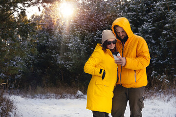 Young couple, man and woman in yellow winter jackets. A man shows on a smartphone, uses a technological gadget. They smile, have a good mood. Pine forest in the background. Sunset. Copy space