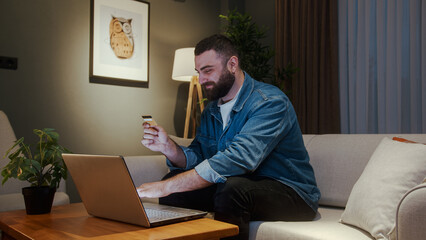 Handsome young adult man sitting on sofa while enter credit card number on laptop for makes secure...