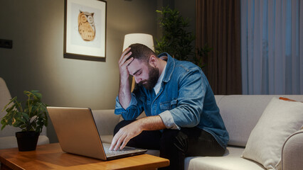 Young man sitting on sofa at home while using laptop having anxiety and stress. Upset young man reacting to loss, bad news on laptop - obrazy, fototapety, plakaty