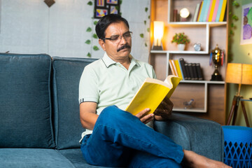 Middle aged man with eyeglasses reading book or novel while sitting on sofa at home - concept of knowledge, hobbies and weekend lifestyle