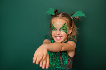 A small, pleasant, cheerful Girl with a painted face with a dragon image laughs looking at the camera. Studio portrait of a child on a green background
