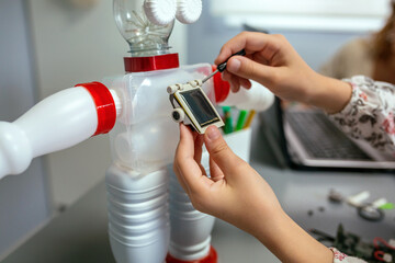 Unrecognizable female student screwing solar panel on recycled toy robot made with plastic packages...