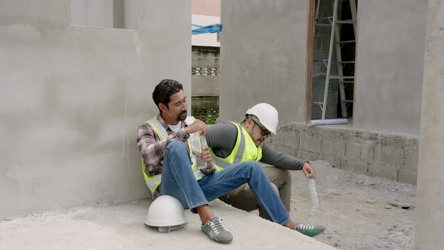 Two Asian male construction engineers wearing hard hats, reflective vests, for safety when working in construction zone, sit rest from construction work, drink water relieve their fatigue.