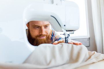 handsome redhaired man with long beard sews at a sewing machine at home studio