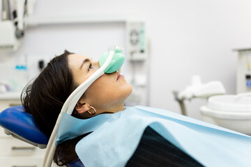 An adult woman sits in a dentist's office wearing a nasal mask to inhale nitrous oxide. Dentist fear concept. Feeling of relaxation with laughing gas. Visit a dentist with relaxation. - obrazy, fototapety, plakaty