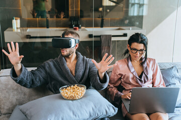Bearded young man in bath robe sitting in bed using virtual reality headset while his wife works using laptop. Domestic leisure, gaming, emote work. Guy playing games home. Woman with computer. - obrazy, fototapety, plakaty