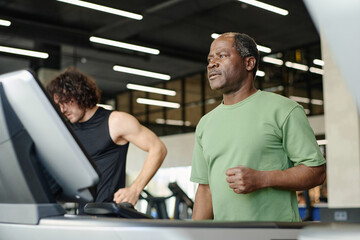 Fototapeta na wymiar Elderly black man standing on treadmill at gym with caucasian gym goer working out on background