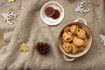 Fototapeta na wymiar Bowl of cookies, cup of tea, dry oranges, pine cones, book, reading glasses and various neutral Christmas decorations on soft beige blanket. Cozy Christmas hygge. Top view.