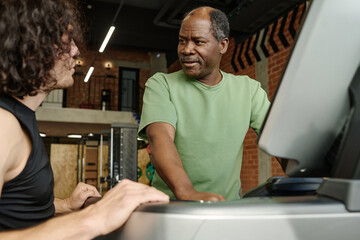 Elderly black man standing on treadmill at gym talking to young caucasian guy standing next to him