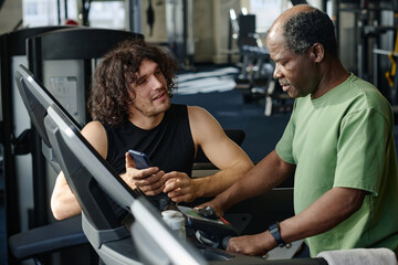Young caucasian sportsman talking to elderly black gym goer, standing on treadmill at fitness center
